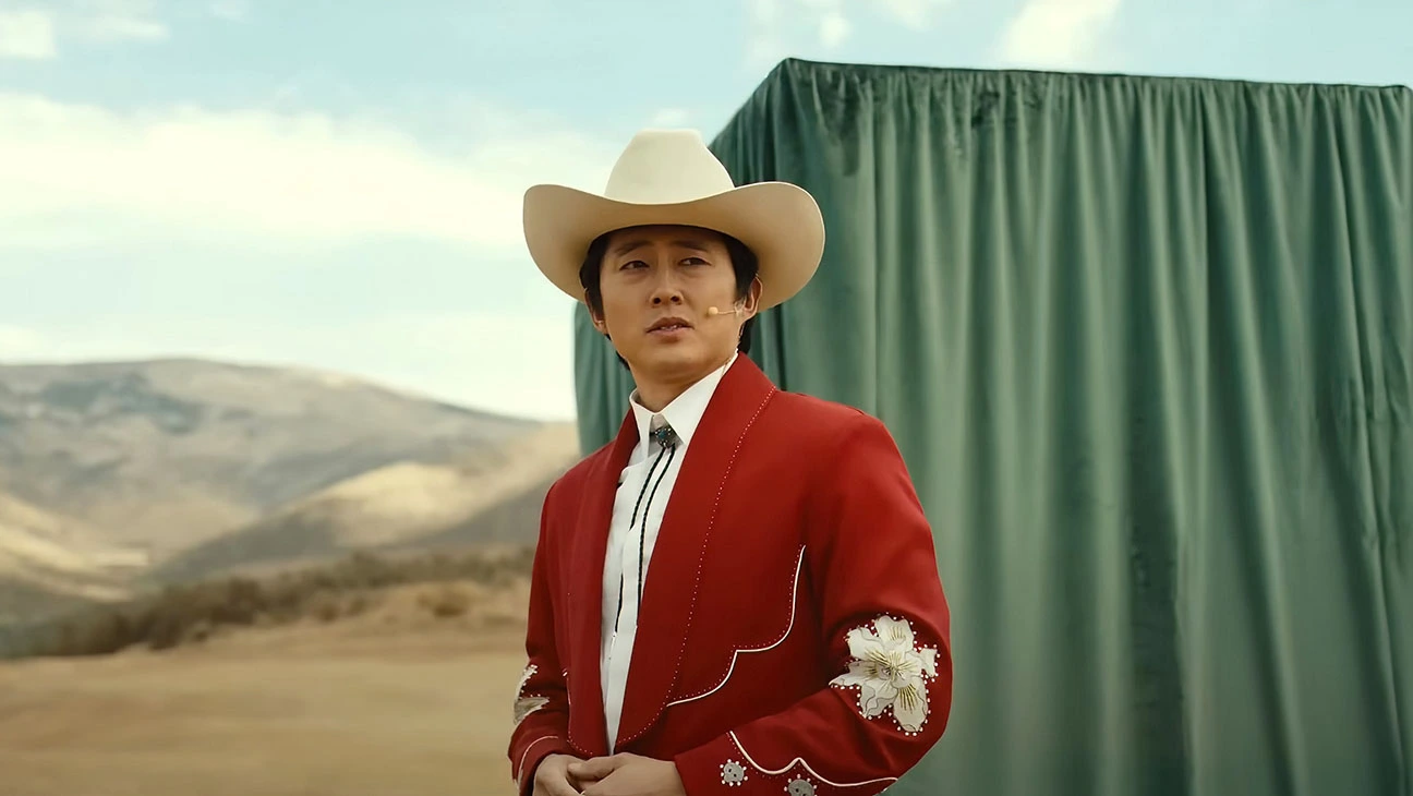 An image of Steven Yeun as Jupe in a red suit and cowboy hat standing in front of a green curtain