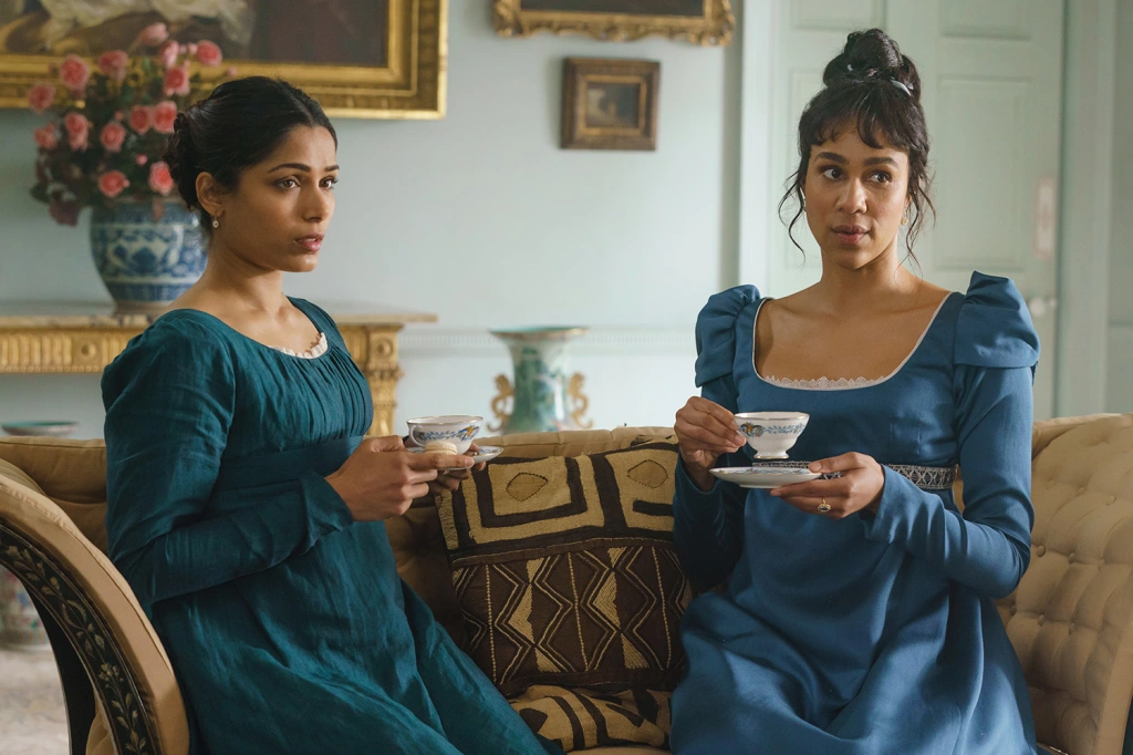 Two women sip tea on a couch wearing matching blue dresses. Left: Freida Pinto as Selina. Right: Zawe Ashton as Julia