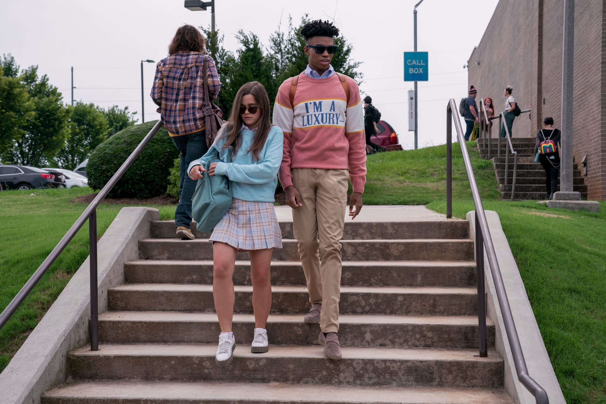 Ben, wearing sunglasses and dressed in a sweater that says "I am a luxury", walks down the steps outside next to Jules, also wearing sunglasses