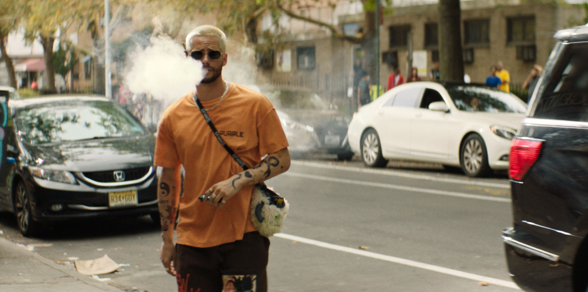 An image of Dylan O'Brien as Colin dressed in a baggy orange shirt, blowing a lot of smoke out on the street