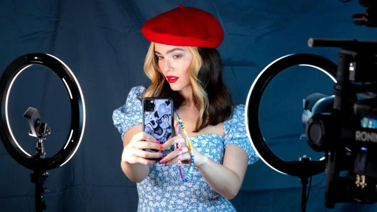 Zoey Deutch as Danni wears a red beret and takes selfies behind ring lights against a blue drape