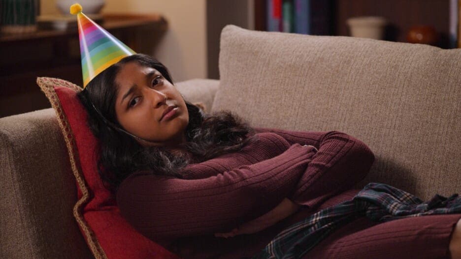 An image of Devi wearing a cone party hat, laying down on a couch, arms crossed, frowning. 