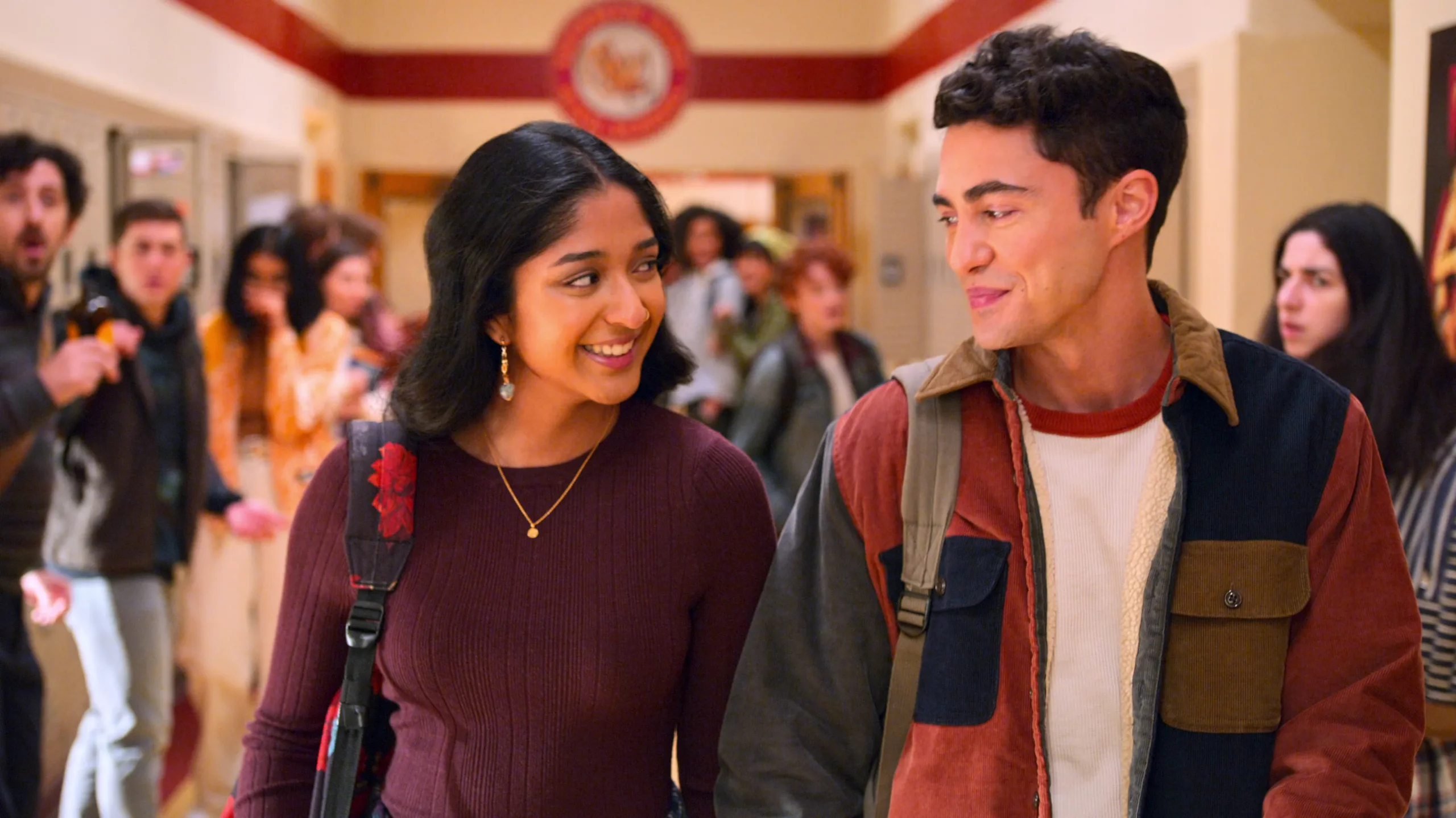 Paxton and Devi walk hand-in-hand, smiling at each other, down a crowded high school hallway