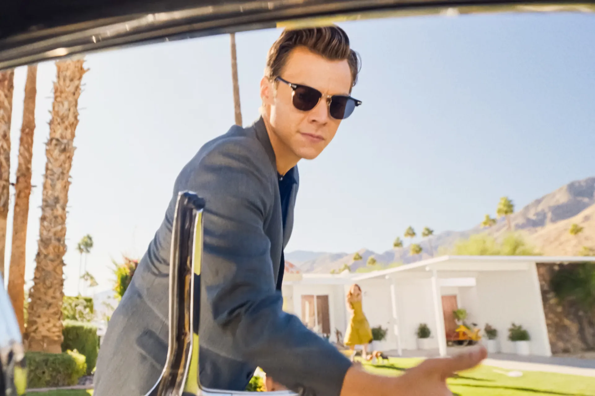 A still from Don't Worry Darling of Harry Styles as Jack dressed in a suit and sunglasses offering his hand to someone as they step out of a car into a sunny 1950s suburb