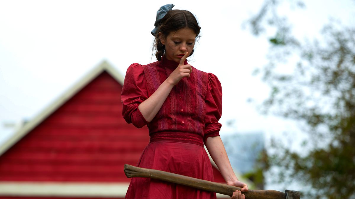 A still from Pearl of Mia Goth as Pearl dressed in a red dress holding an axe and shushing someone