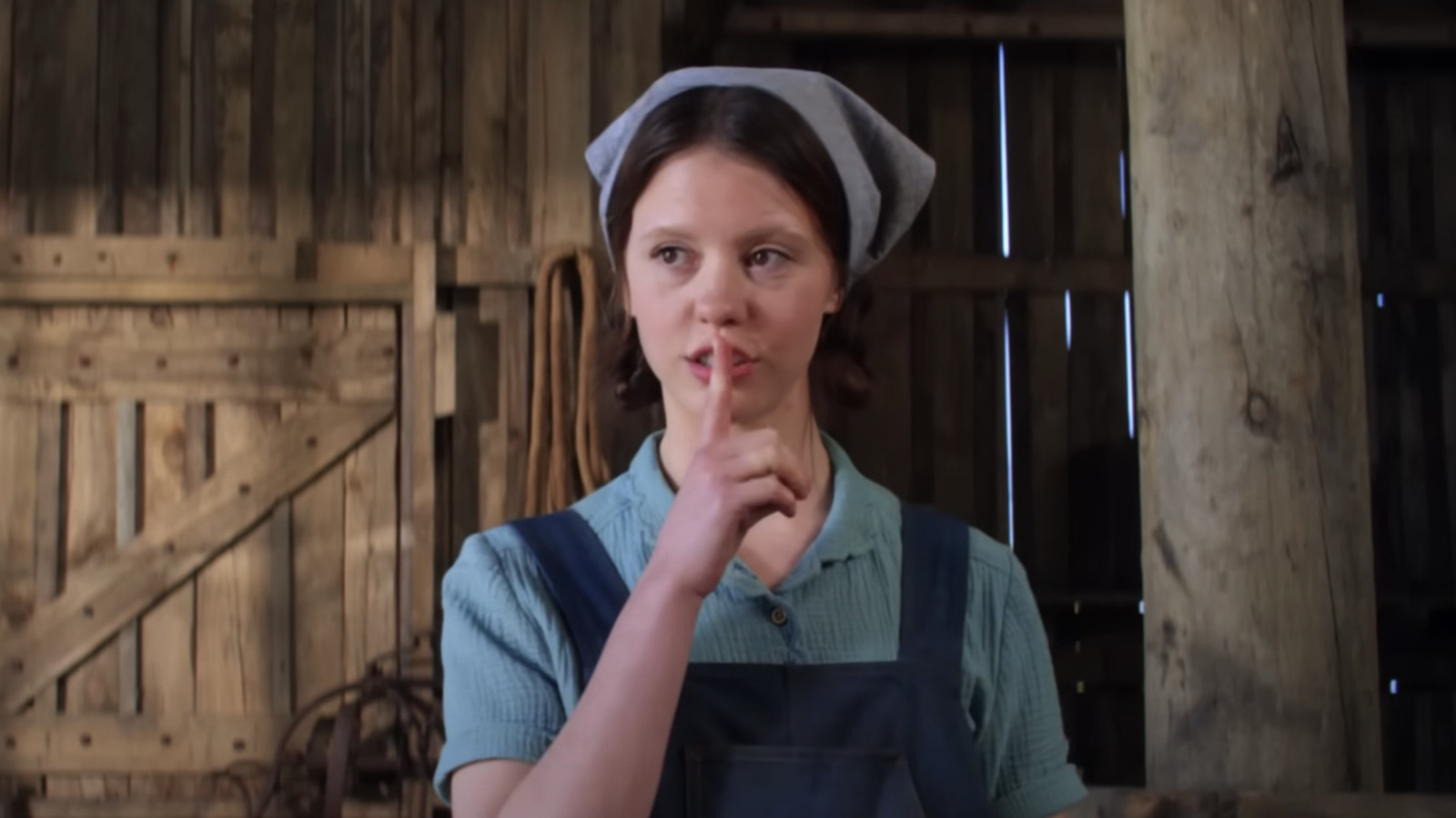 A still from Pearl of Mia Goth as Pearl dressed in farming clothes, putting her finger to her mouth in a shushing gesture