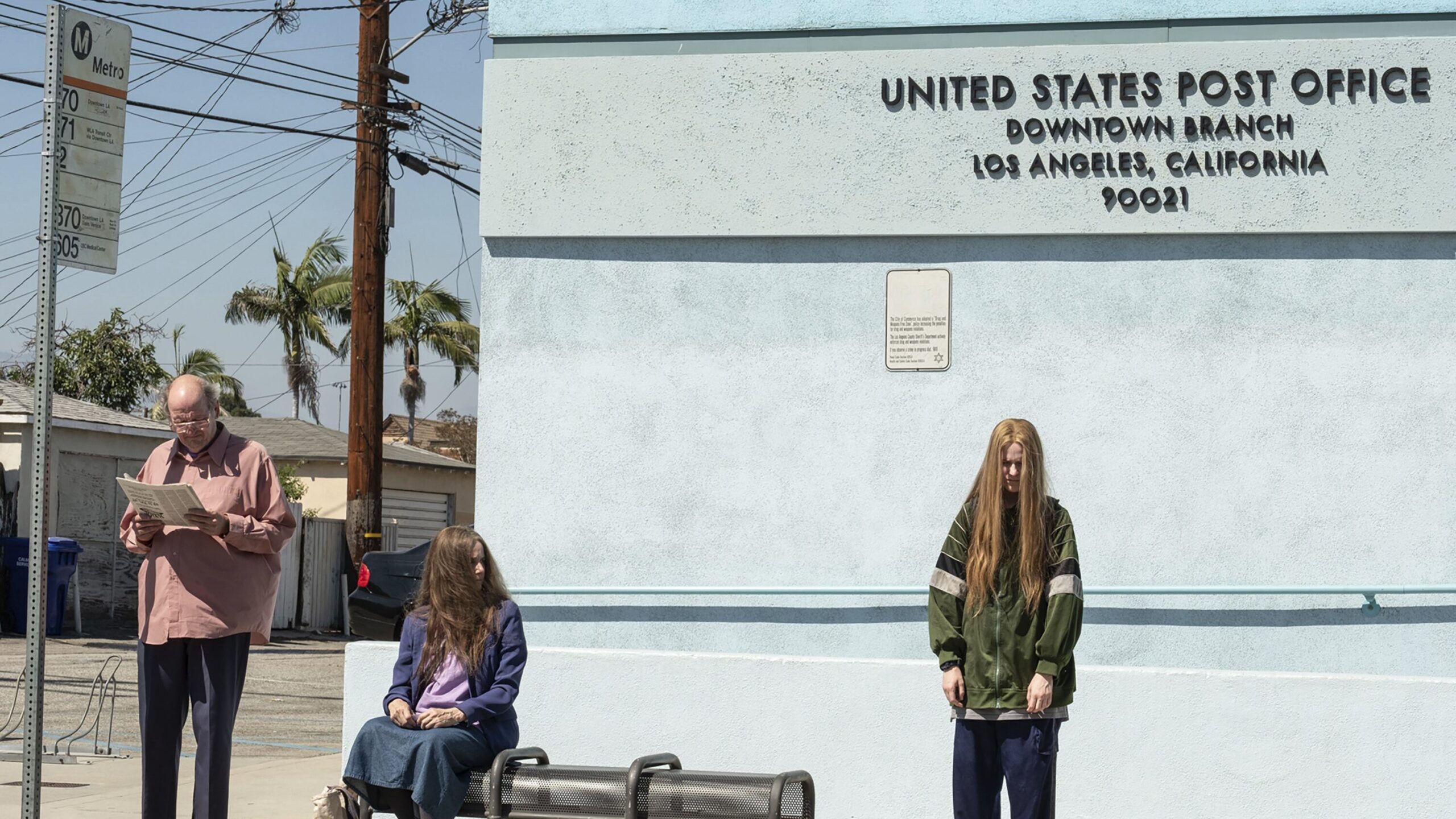 Robert, Theresa, and Old Dolio wait at a bus stop. Old Dolio stands apart from her parents and appears upset.
